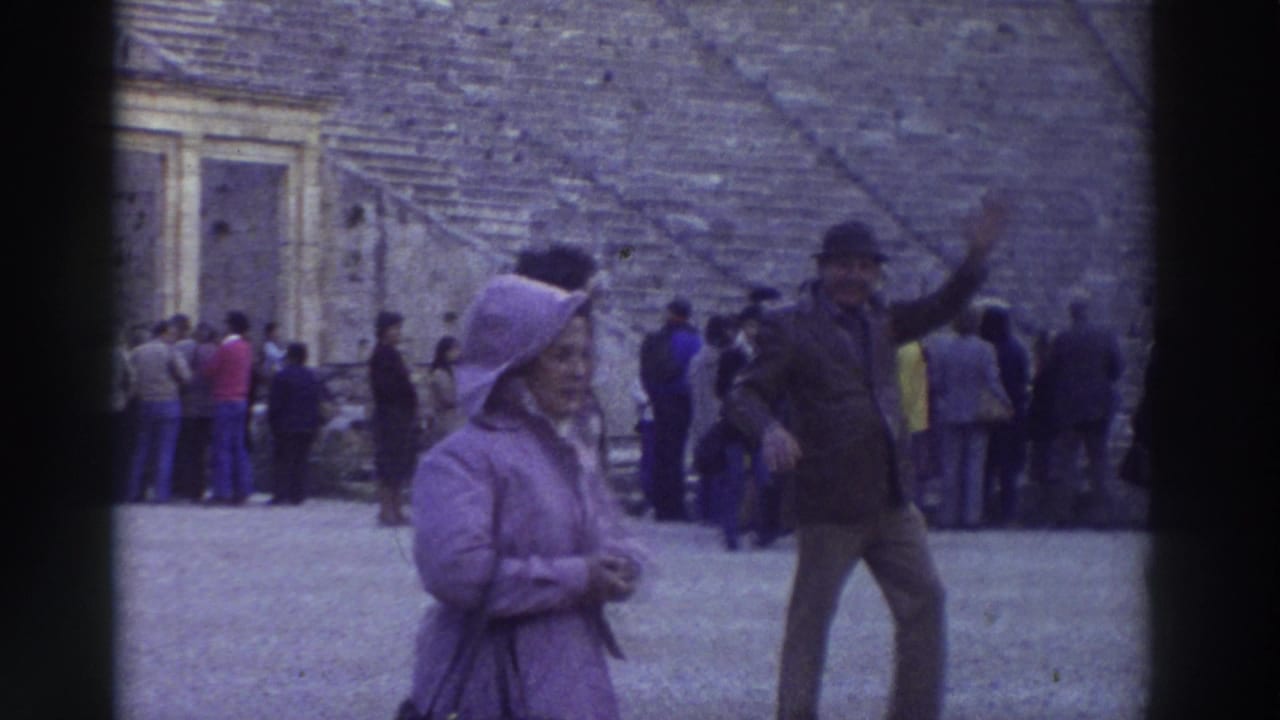 1976 home-movie still: woman in purple hooded coat and waving man in dark cap stand at the floor of an ancient Greek theater, Greece; other tourists crowd the background.