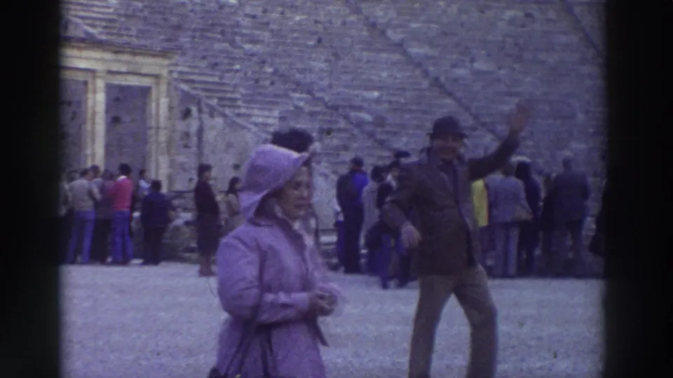 1976 home-movie still: woman in purple hooded coat and waving man in dark cap stand at the floor of an ancient Greek theater, Greece; other tourists crowd the background.