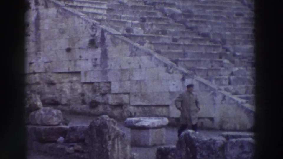 1976 home-movie still: expansive ancient stone theater cavea with tiered seating filling the frame, Greece; a lone visitor stands to the right on the stone steps.