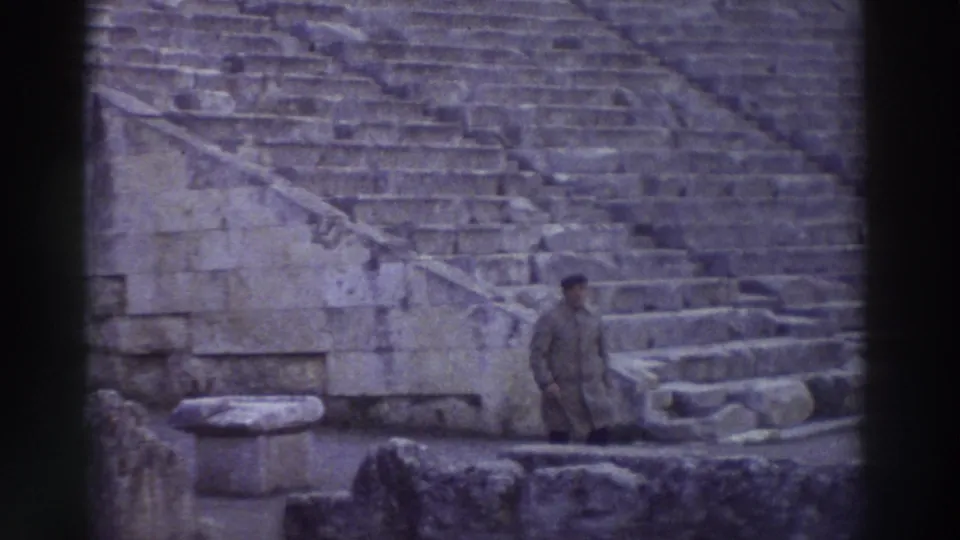 1976 home-movie still: man in dark coat pauses at the lowest rows of an ancient Greek theater's stone cavea, Greece; tiered seating rises steeply behind him.