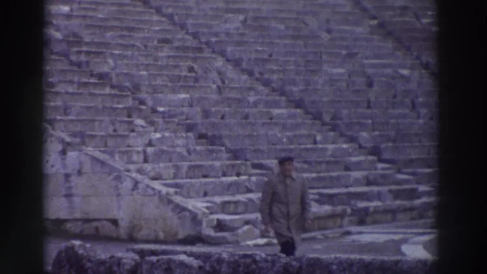 1976 home-movie still: man in dark coat and cap stands alone at the base of an ancient Greek theater's cavea, Greece; stone seating tiers rise steeply in the background.
