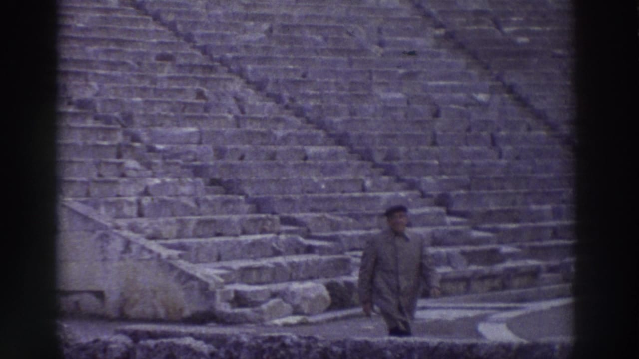 A man in a long coat and cap stands on the stone seating tiers of an ancient Greek amphitheater, captured on colour-shifted 8mm home film, circa 1976.