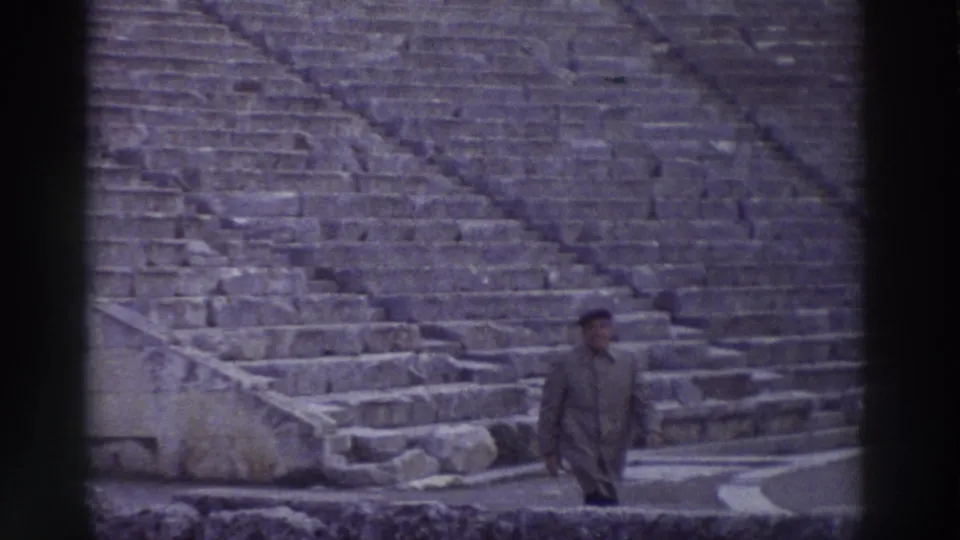 A man in a long coat and cap stands on the stone seating tiers of an ancient Greek amphitheater, captured on colour-shifted 8mm home film, circa 1976.