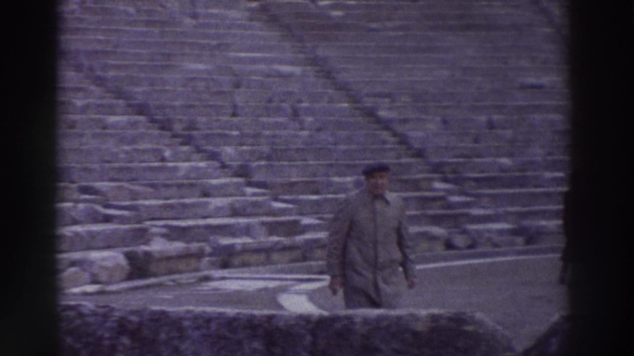The same man in coat and cap moves across the stone seating area of an ancient amphitheater in Greece, 8mm home film circa 1976.