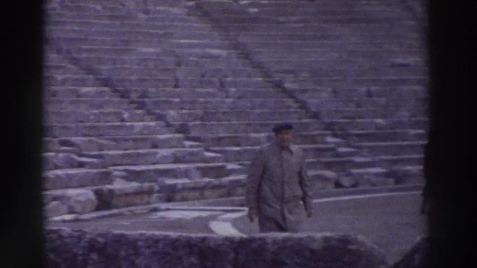 The same man in coat and cap moves across the stone seating area of an ancient amphitheater in Greece, 8mm home film circa 1976.