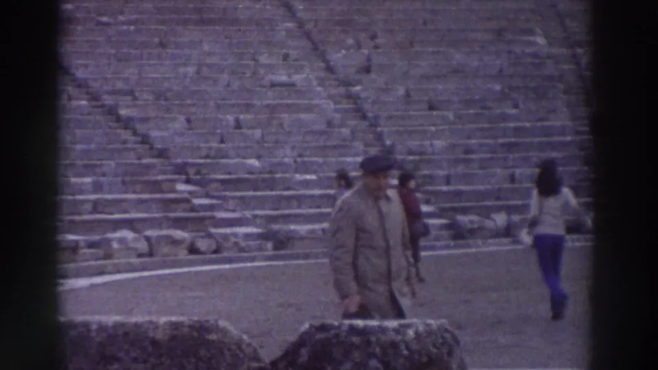 A man in a long coat walks toward the camera on the stage floor of an ancient Greek amphitheater; other visitors in 1970s clothing visible behind him, circa 1976.