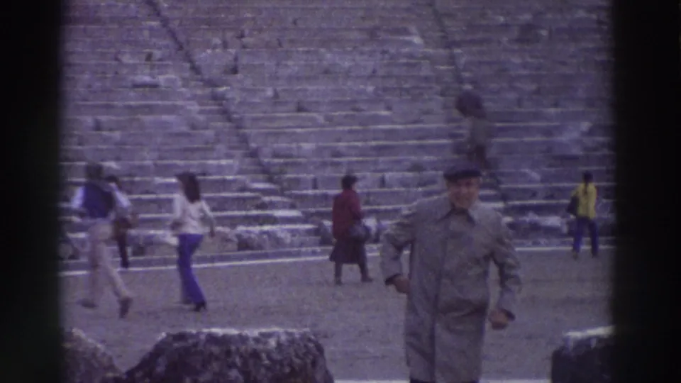 Close view of a man in a coat near the camera with multiple visitors scattered behind him on the stage of an ancient amphitheater in Greece, 8mm home film circa 1976.