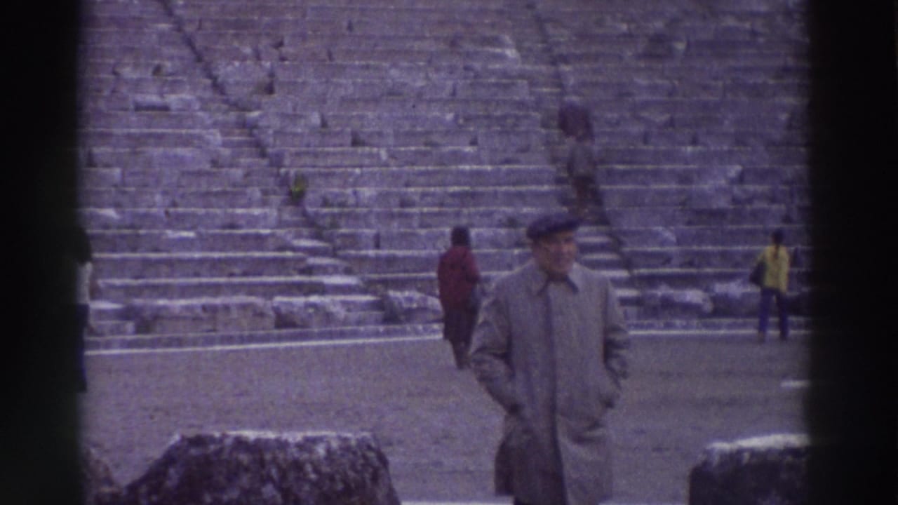 Man in trench coat and flat cap stands before a vast tiered stone amphitheater, two other visitors visible on the steps.