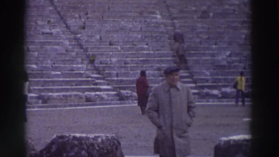 1976 Greece: man in trench coat and beret at a large ancient stone amphitheater, other tourists visible on the tiered stone steps.