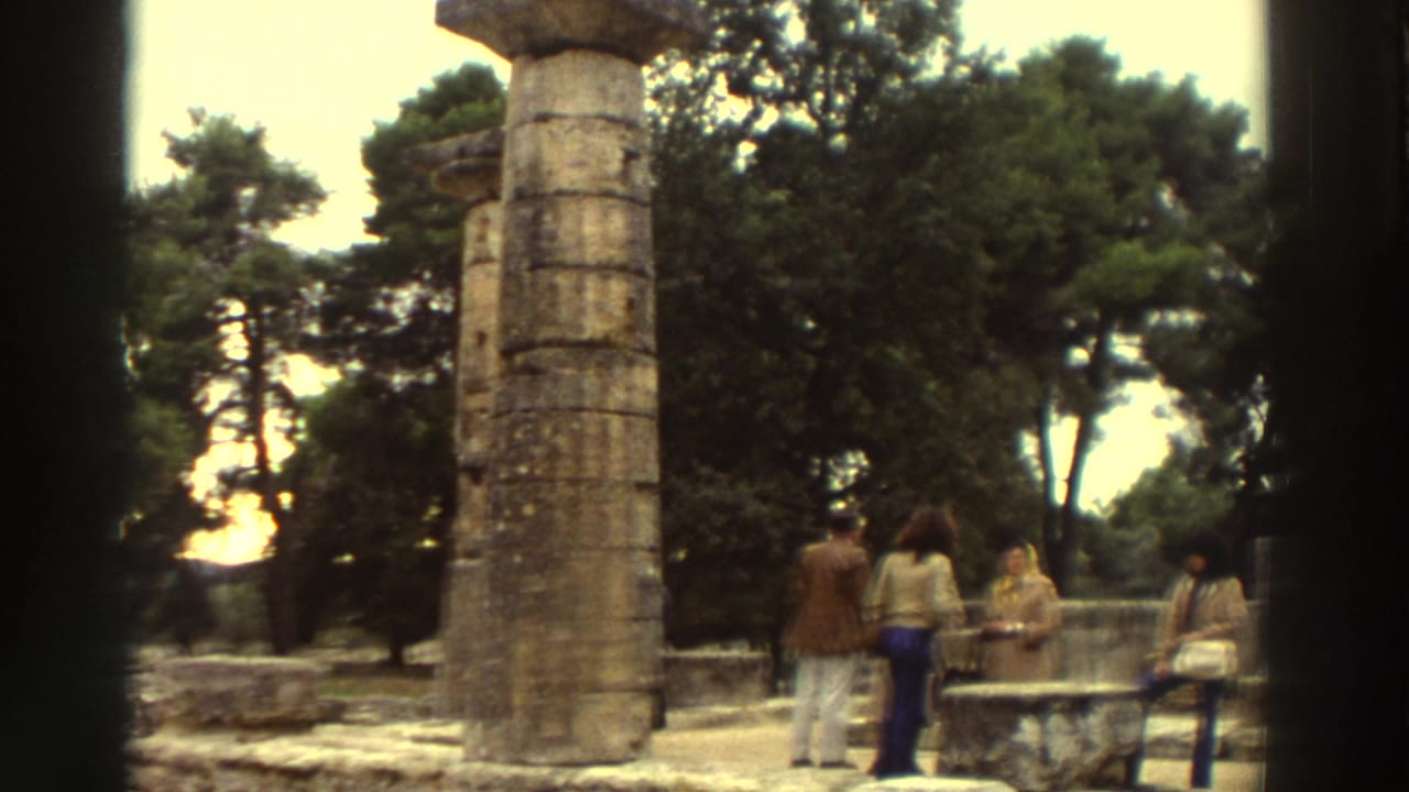 Tall ancient drum-stacked stone column dominates left frame; small tourist group in 1970s clothing stands at right among ruins.