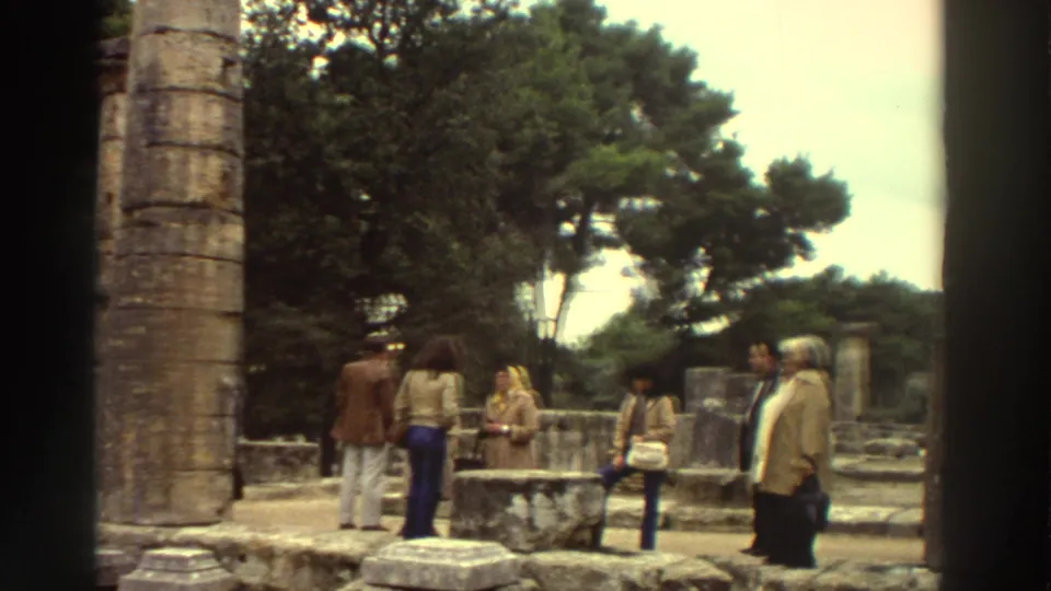 1976 Greece: tourist group in 1970s casual wear including rust jacket and wide-leg trousers stand among stone ruins, ancient column at left.