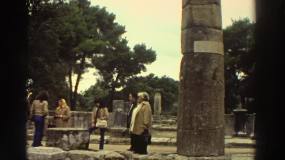1976 Greece: tourist group in 1970s attire near large ancient column, woman with handbag, additional column fragments and ruins in background.