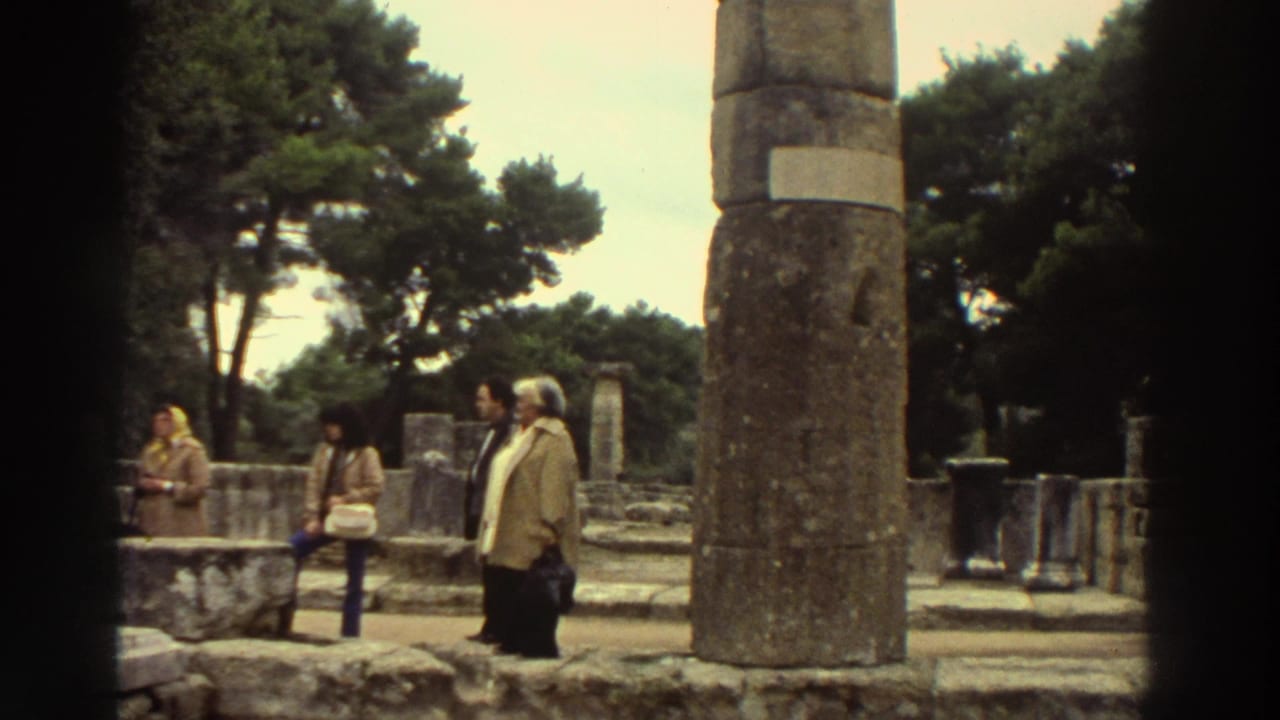 Wide establishing shot of tourists in 1970s dress exploring ancient ruins at a Greek archaeological site, circa 1976, with weathered stone columns and pine trees.