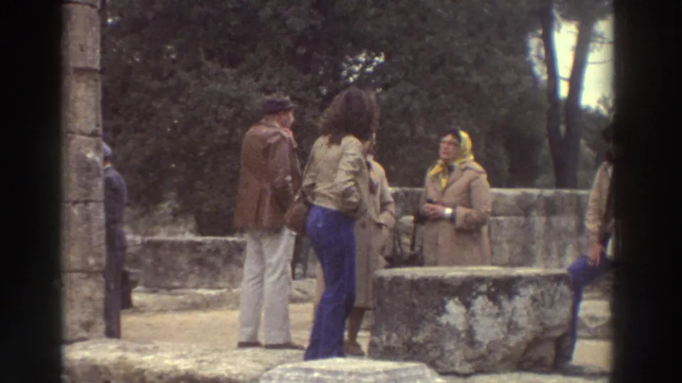 Group of tourists in 1970s attire gathered near stone steps at a Greek archaeological site, circa 1976; man in brown jacket and woman in yellow headscarf visible.