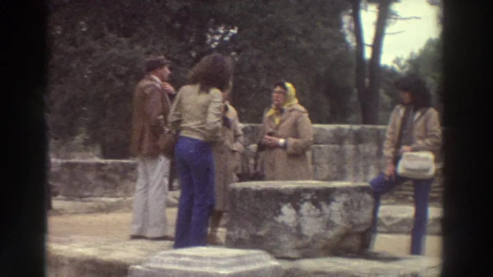Tourists in 1970s dress near ancient stone ruins in Greece, circa 1976; woman in yellow headscarf and beige coat prominent on right with shoulder bag.