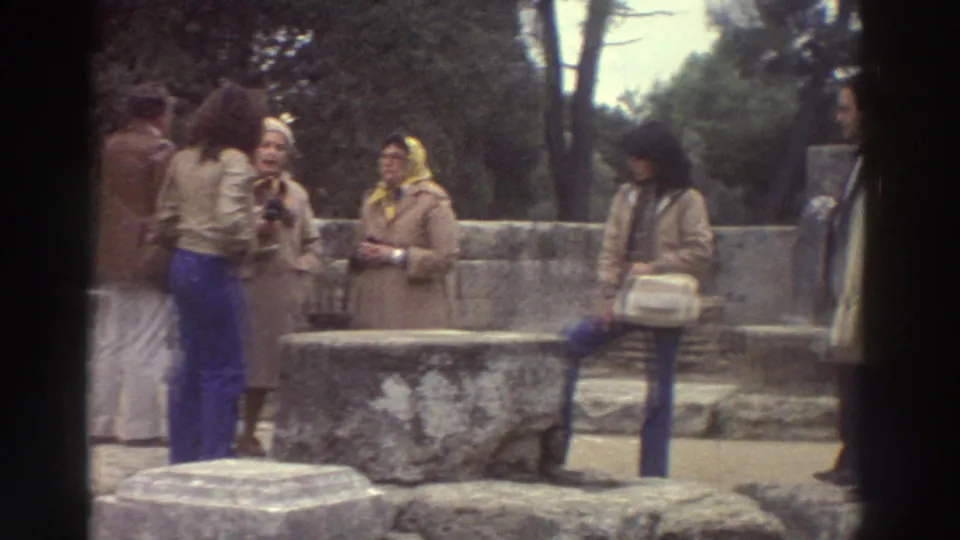 Three or four tourists in 1970s earth-tone coats, one wearing a yellow headscarf, standing near weathered stone steps at a Greek archaeological site, circa 1976.