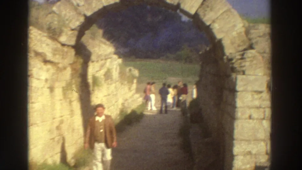 Tourist in a brown jacket walks through a monumental stone vaulted passage at a Greek archaeological site, circa 1976, with a group of visitors in the background.
