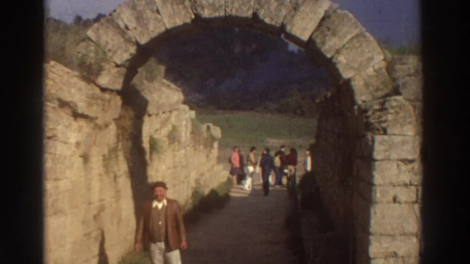 Tourist in a brown jacket moves through a massive ancient stone vaulted archway at a Greek ruin site, circa 1976, other visitors visible in the sunlit passage beyond.