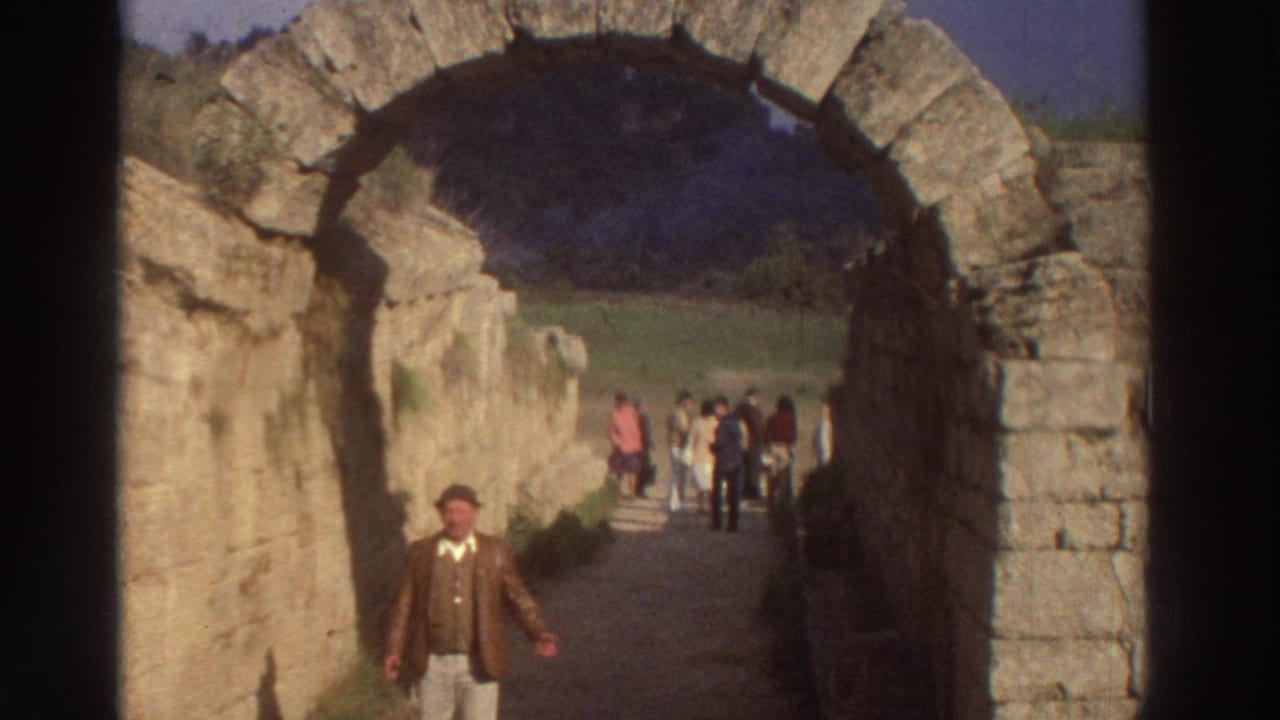 1976 8mm footage of a man in 1970s dress approaching through an ancient cut-stone barrel arch at a Greek archaeological site, tourists grouped in the sunlit background.