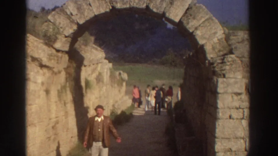 1976 8mm footage of a man in 1970s dress approaching through an ancient cut-stone barrel arch at a Greek archaeological site, tourists grouped in the sunlit background.