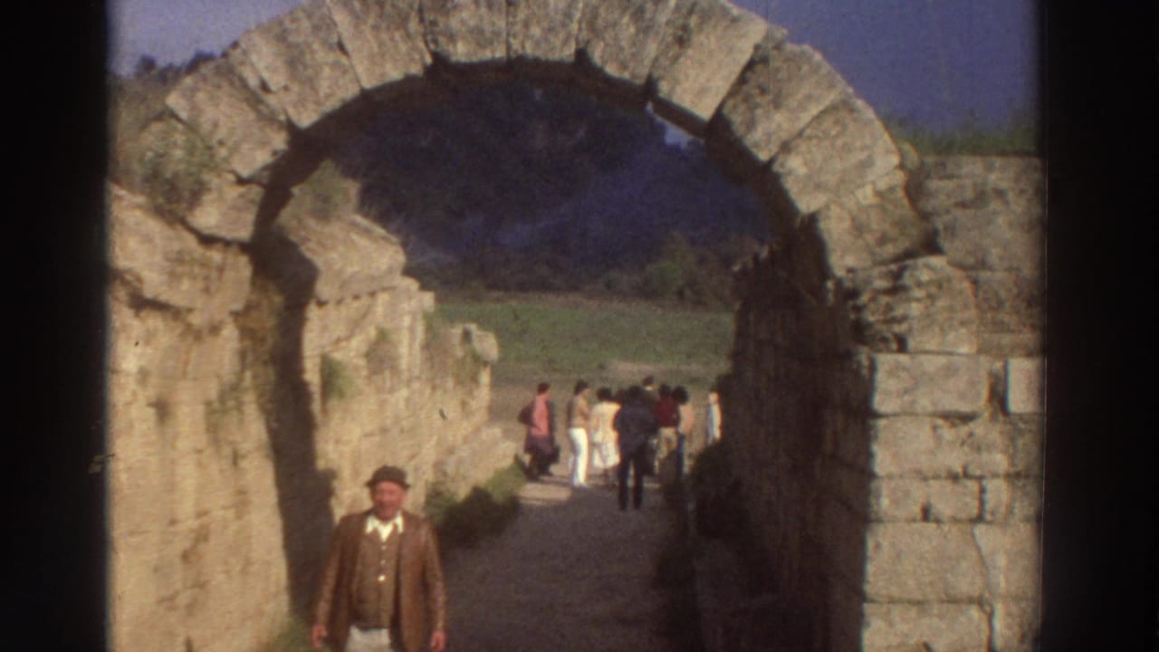 Man in brown jacket and cap now closer to camera, framed within a large cut-stone arch; tourists walking along a path visible through the arch behind.