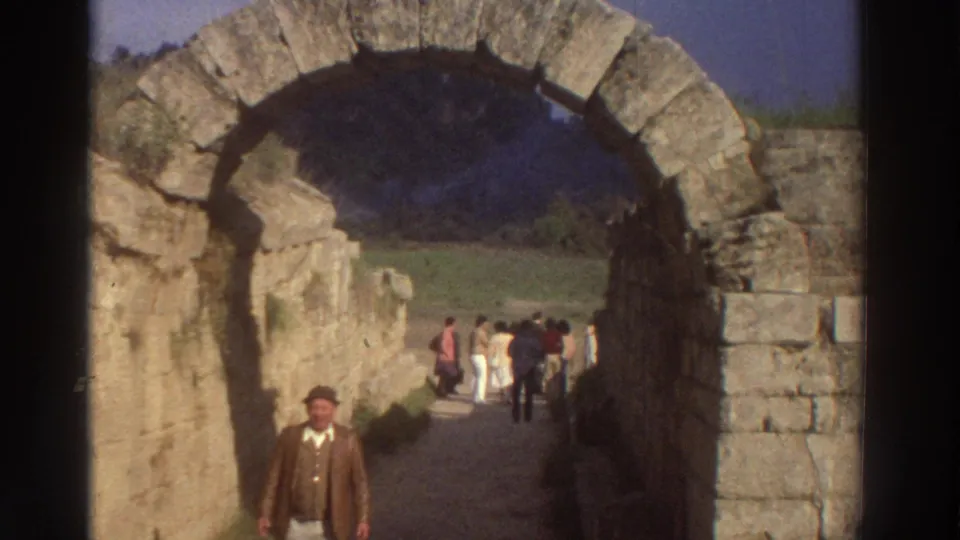 8mm frame from 1976 Greece showing a man in 1970s dress nearer to camera within an ancient stone archway, tourists strolling in bright background beyond.
