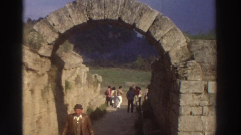 8mm frame from 1976 Greece: man in brown jacket and flat cap nearly at camera, ancient stone arch framing, crowd of 1970s-dressed tourists visible on path behind.