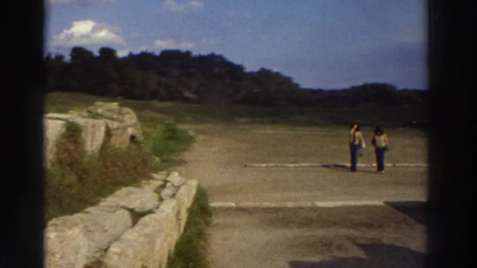 8mm frame from 1976 Greece: wide view of what may be an ancient open precinct or stadium, a few tourists as small distant figures on a broad paved area, rolling hills and blue sky behind.