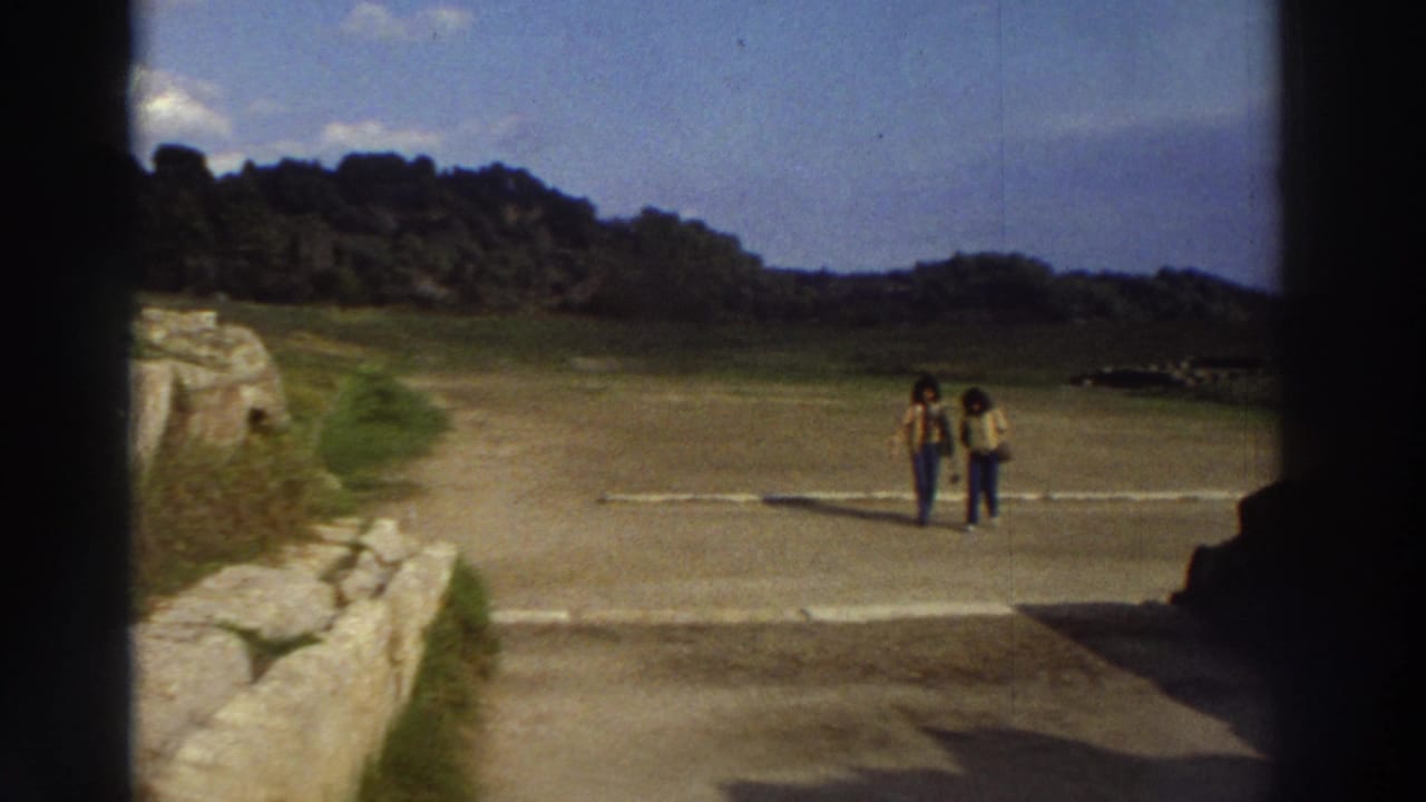 Two women wearing bell-bottom jeans and casual tops walk toward camera at a Greek archaeological site circa 1976; ancient low stone wall in foreground.