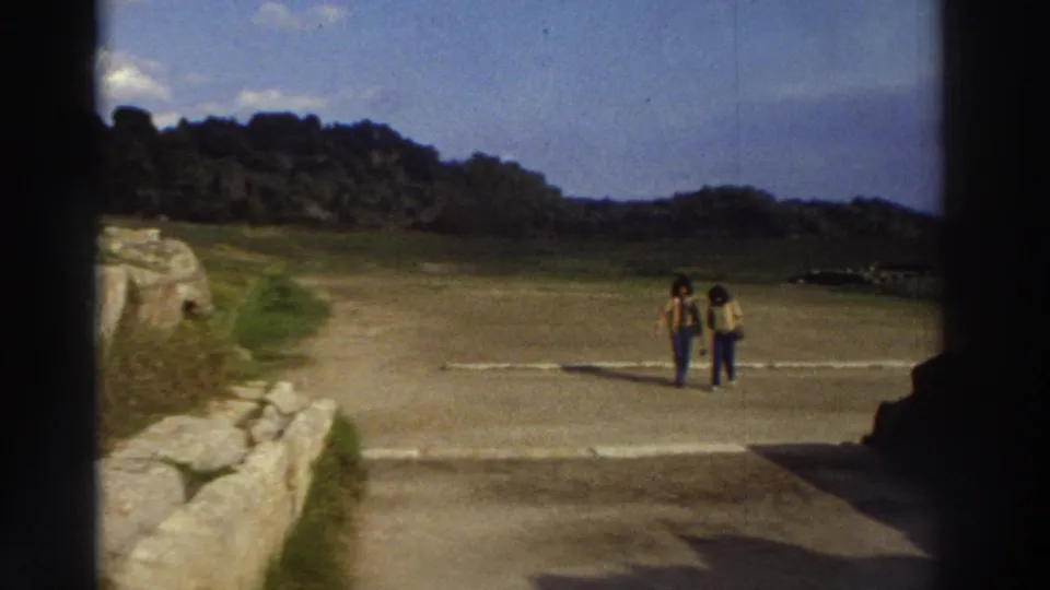 Two women wearing bell-bottom jeans and casual tops walk toward camera at a Greek archaeological site circa 1976; ancient low stone wall in foreground.