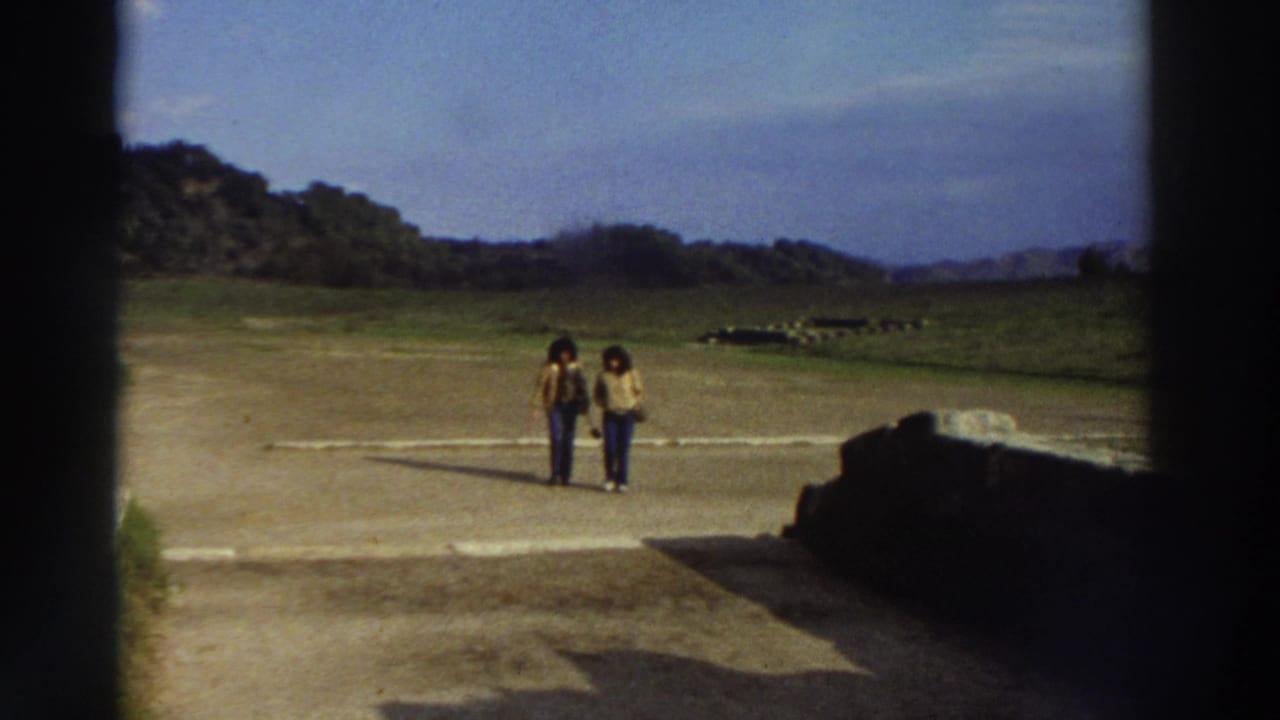 Two women in casual 1970s attire walking on a path at an open archaeological site, ancient stone ruins visible at left.