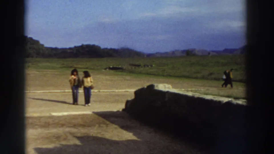Wide view of two women in 1970s clothing crossing open ground at a Greek archaeological site circa 1976; third visitor in distance, green hills beyond.