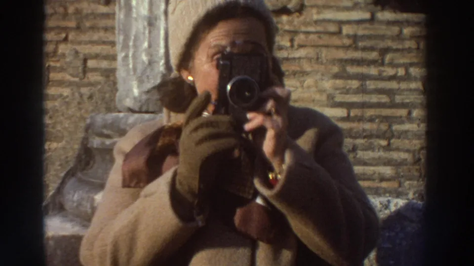 Woman in camel coat and gloves raising a camera to her eye at ancient ruins in Greece, circa 1976, framed between rough stone masonry walls.