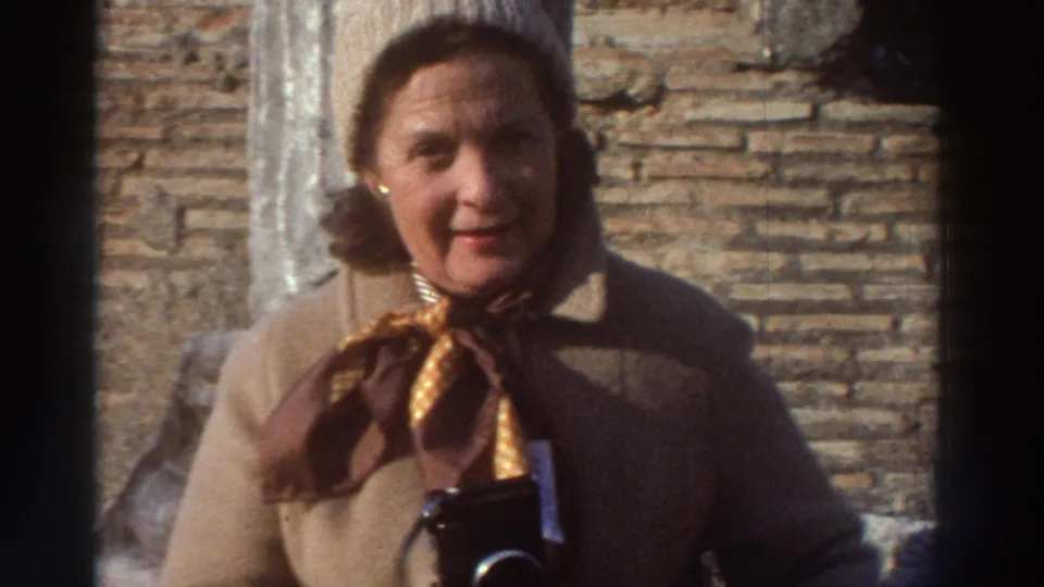 Middle-aged woman in camel coat and patterned gold scarf at Greek ruins, circa 1976, looking toward the camera with a camera held at her side.