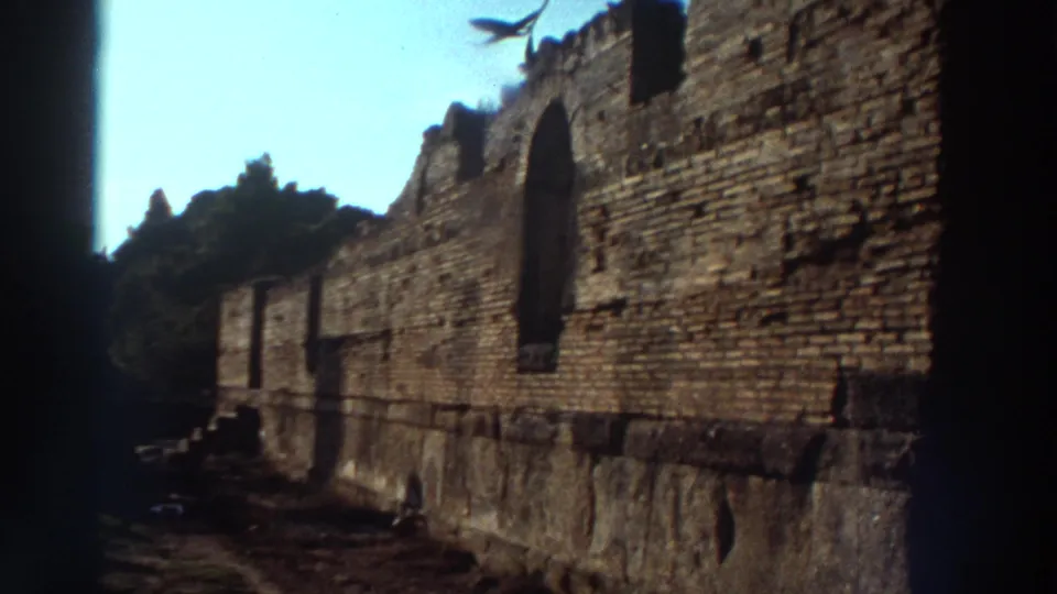 Ancient stone fortification or ruin wall at a Greek archaeological site, circa 1976, shot from ground level looking along the wall toward distant hills and treeline.