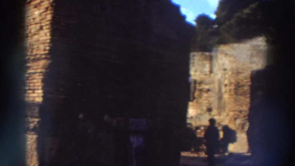 Interior of ancient Greek ruin with silhouetted tourists passing through a stone passageway, trees and blue sky visible above, and a green lens flare on 8mm film, 1976.