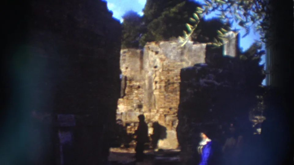 Greek archaeological ruin interior, 1976: visitor in blue clothing stands near low broken stone walls, with Mediterranean trees, possible site signage, and blue sky behind.