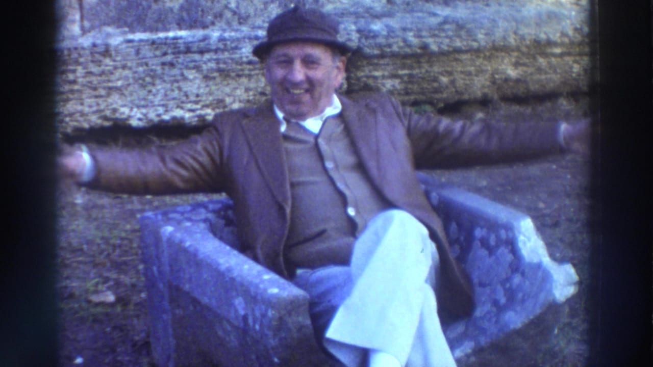Older man in brimmed hat and brown jacket seated on a stone surface, smiling and spreading his arms wide toward the camera.