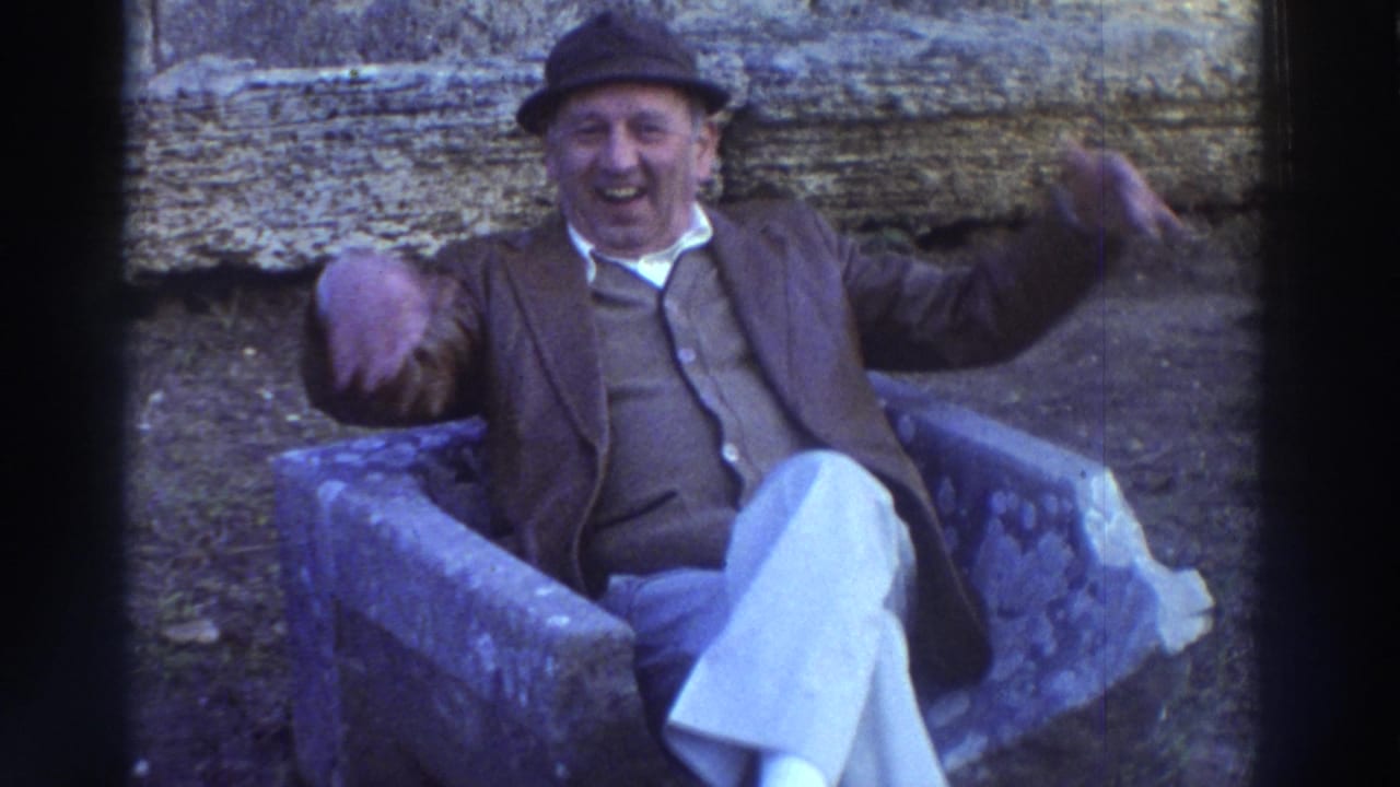 The same older man in 1970s attire continues an animated wave and laugh toward the camera, seated on a stone bench in Greece circa 1976.