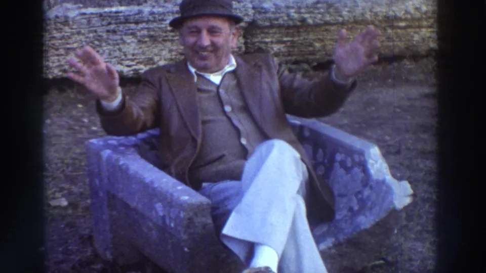 Closer view of the older man in 1970s attire leaning forward with both hands raised in an expressive wave, seated on a stone bench in Greece circa 1976.