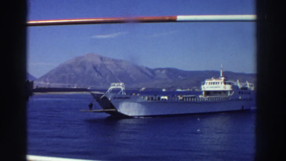 Large white passenger ferry in a Greek harbor circa 1976, with a rocky mountain headland in the background, calm blue water, and a foreground railing visible at the top of the frame.