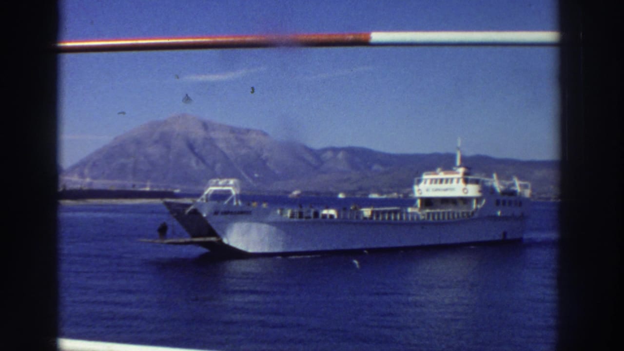 White multi-deck car ferry crossing blue Mediterranean water, large mountain behind, shot from neighboring vessel through a ship railing, Greece, 1976.