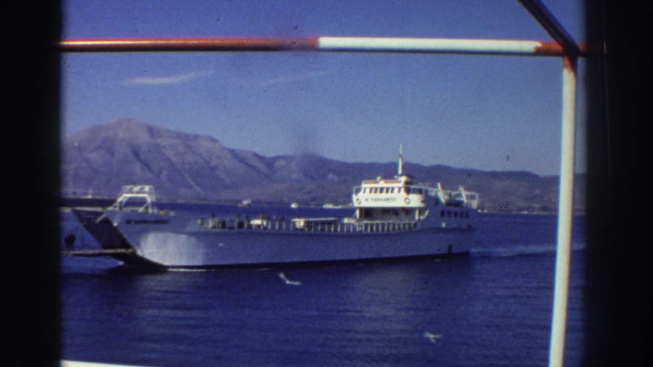 White car ferry on blue sea, mountain ridge behind, vertical ship railing post visible at right edge.
