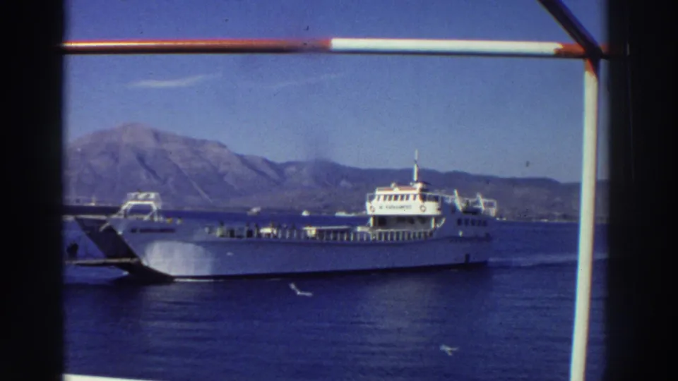 White multi-deck car ferry on blue Mediterranean, mountain ridge behind, vertical railing post at frame edge, Greece, 1976.