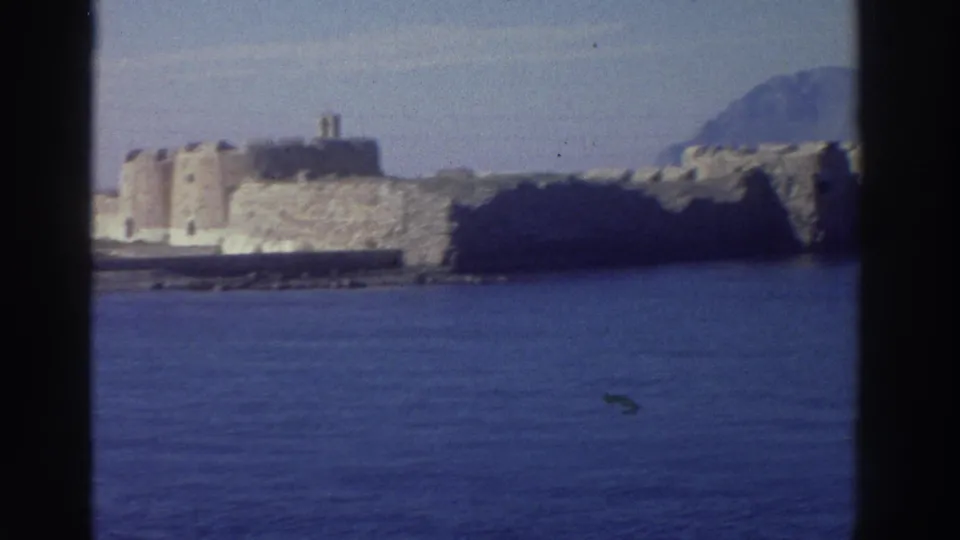 Stone coastal fortress with circular towers on rocky promontory, blue Mediterranean sea, hazy hills to right, Greece, may be 1976.
