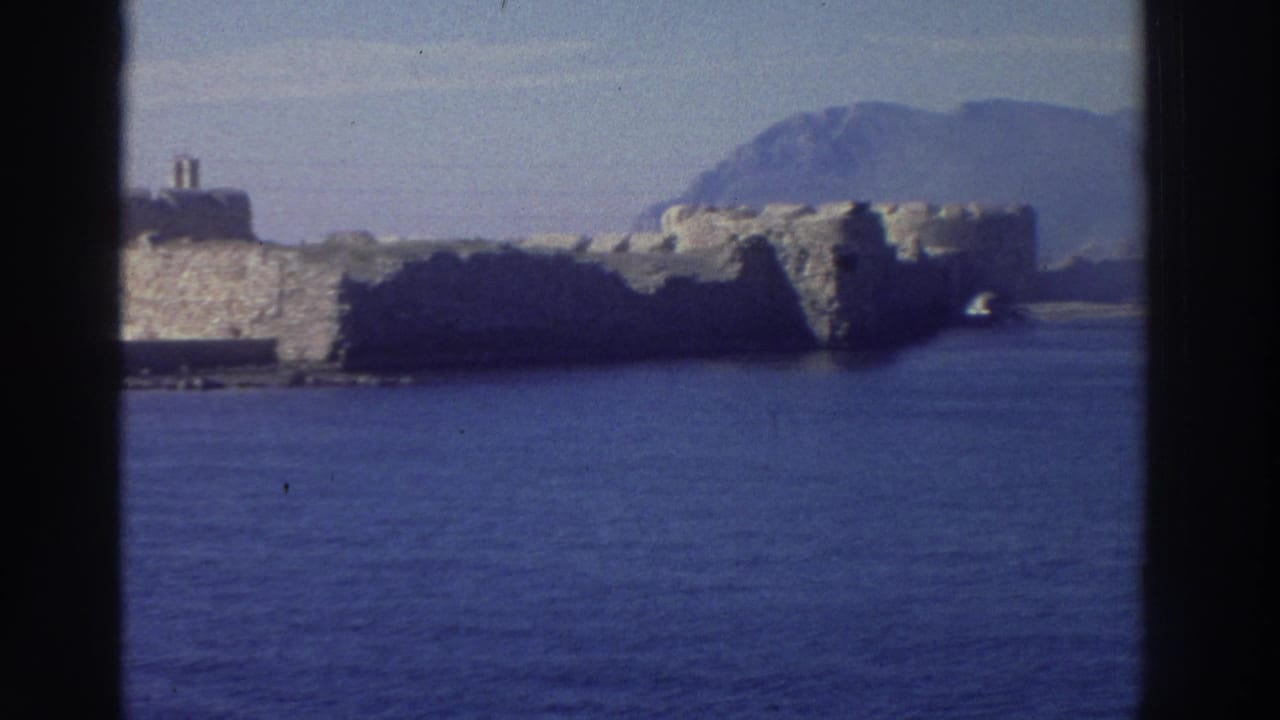 Stone fortress walls rising above the Aegean Sea framed by a boat window, rugged mountain ridgeline visible in background.