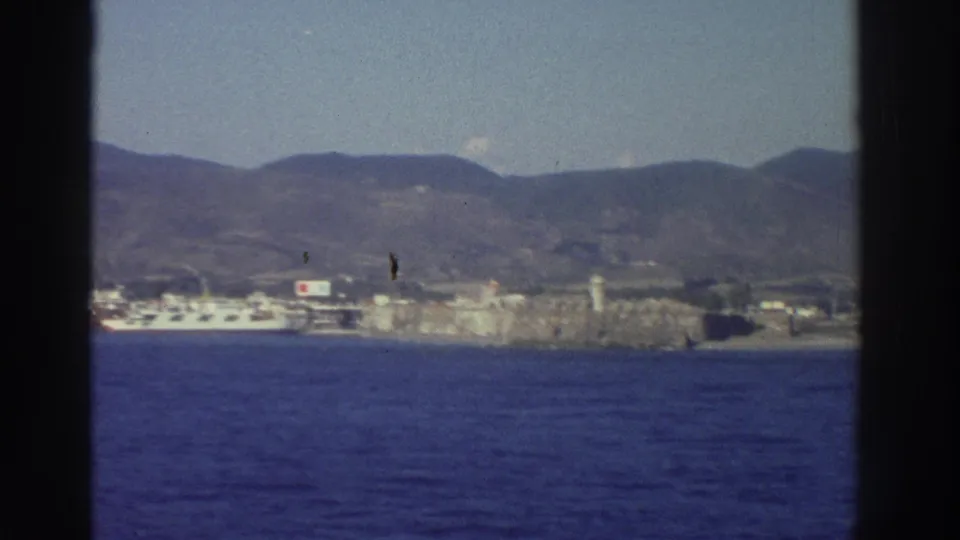8mm footage from Greece, circa 1976, panning to a coastal harbor town with white buildings and mountains; a small marking appears on dock structures and should be reviewed.