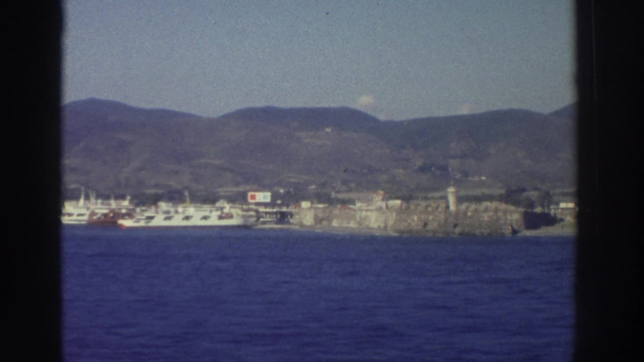 Ship's-eye view of a Greek harbor town, 1976 — white ferry at dock, stone fortress wall, and arid hills behind, filmed on Super 8.