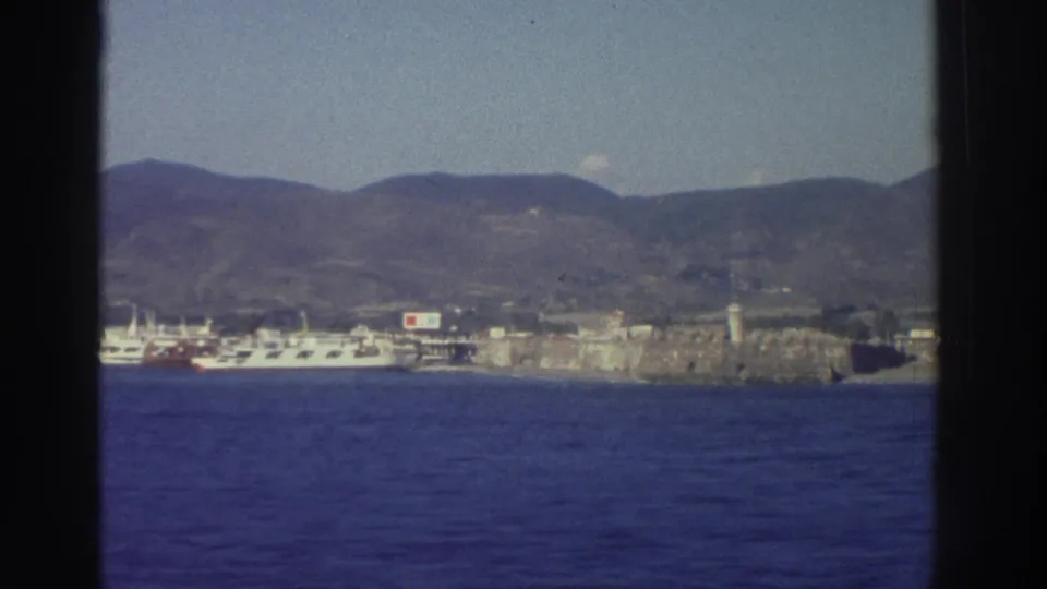 Ship's-eye view of a Greek harbor town, 1976 — white ferry at dock, stone fortress wall, and arid hills behind, filmed on Super 8.