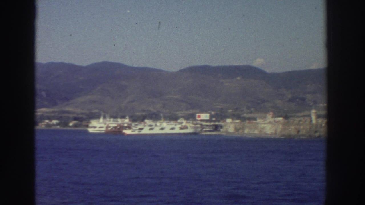 Similar harbor view from the moving ship — white ferry, stone fortification, and arid hillside across blue Aegean water.
